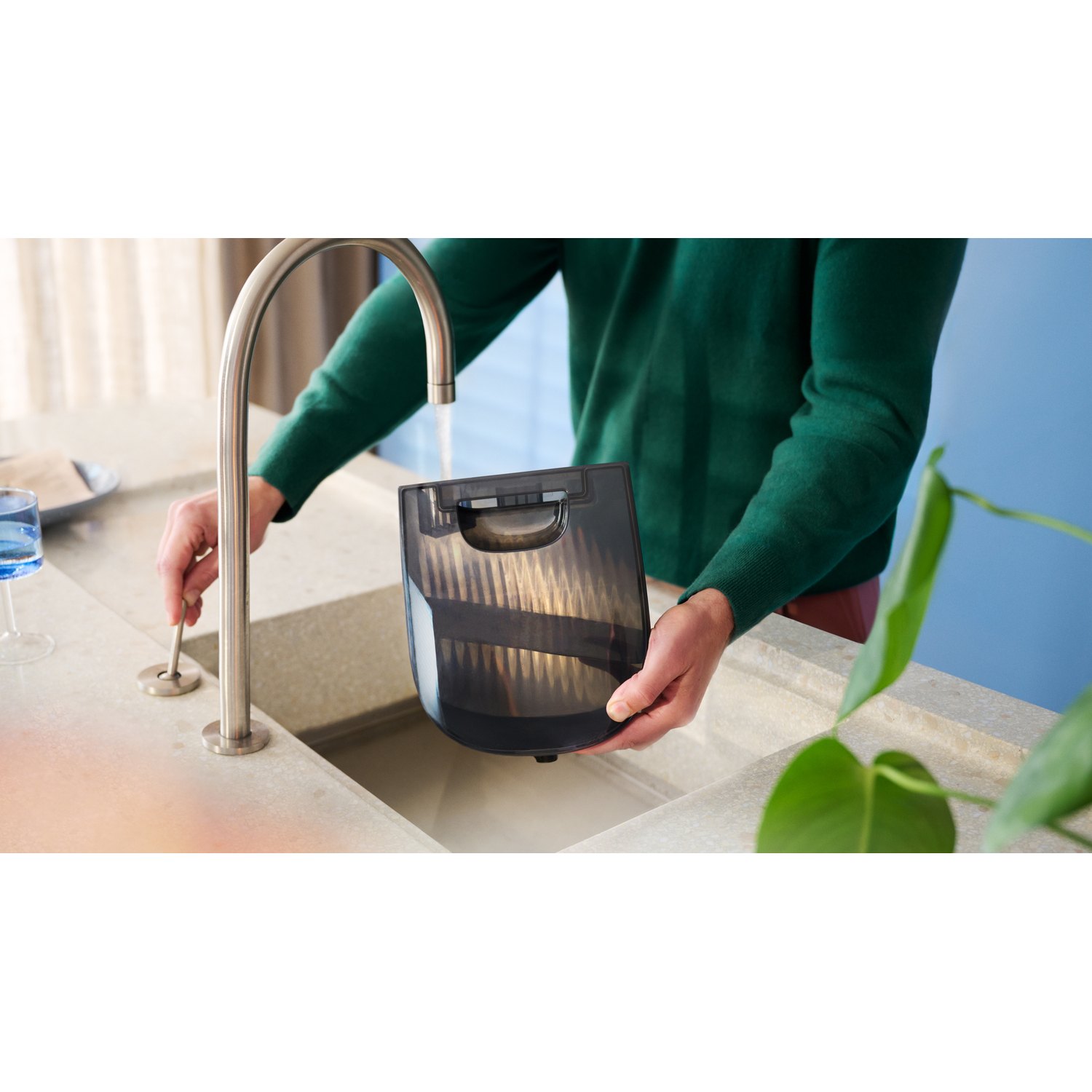 Une personne lave un filtre d'aspirateur dans un évier de cuisine. L'eau coule et une plante est visible.