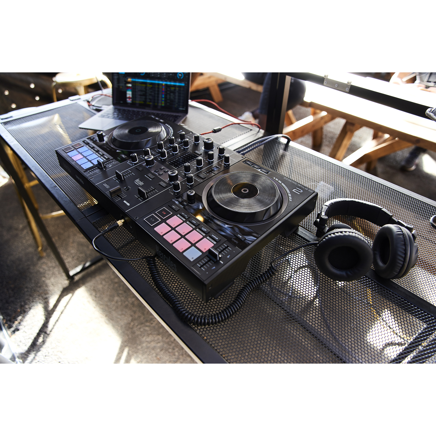 DJ setup on a table. Mixer, laptop, headphones, all black. Metal table, outdoor setting.
