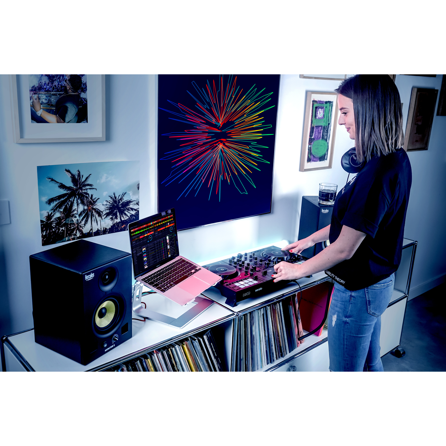 A woman DJs with laptop and controller. Background has art and speakers, with records below. 