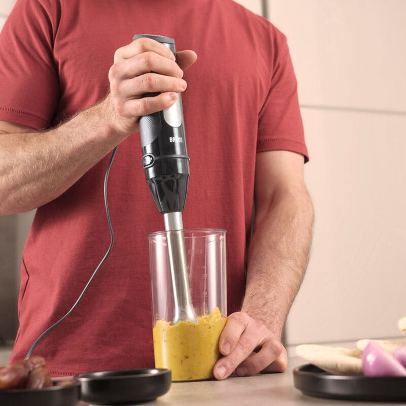 Persona che frulla una miscela gialla con un frullatore a immersione Braun nero in un bicchiere.