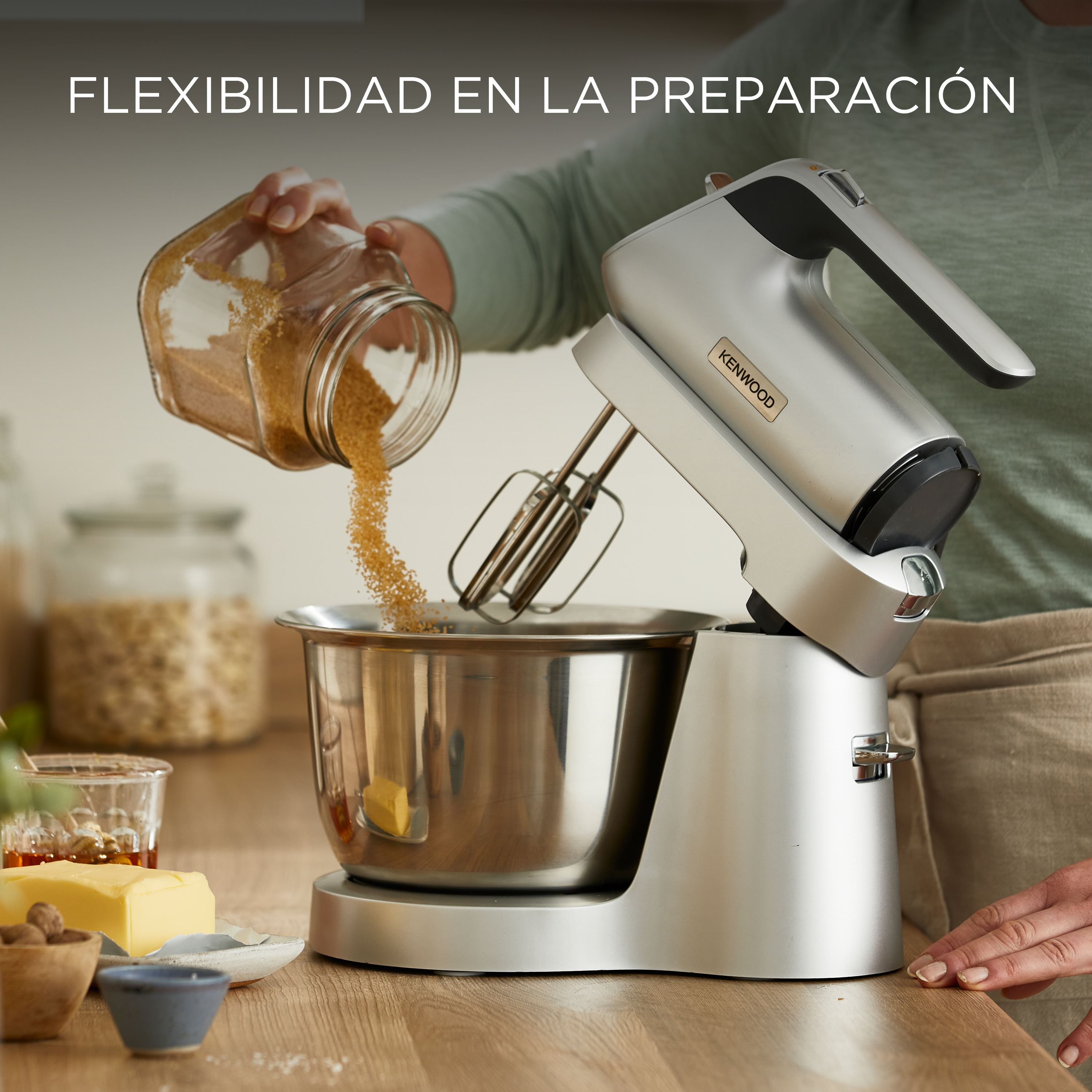 Una persona vertiendo azúcar moreno en una batidora Kenwood plateada, con mantequilla y especias en la mesa.