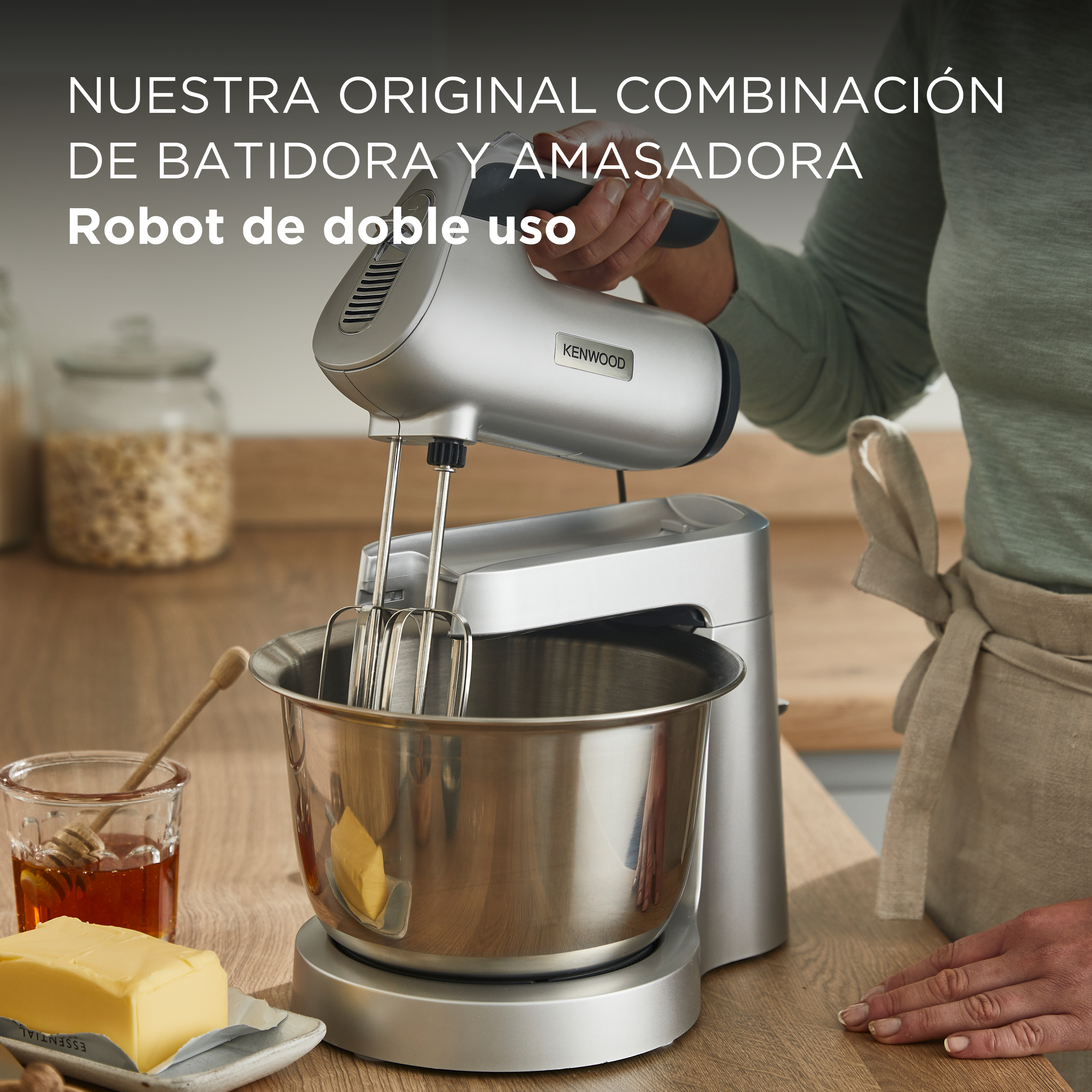 Una batidora Kenwood plateada sobre madera con mantequilla, miel y un vaso. Una persona la sostiene.