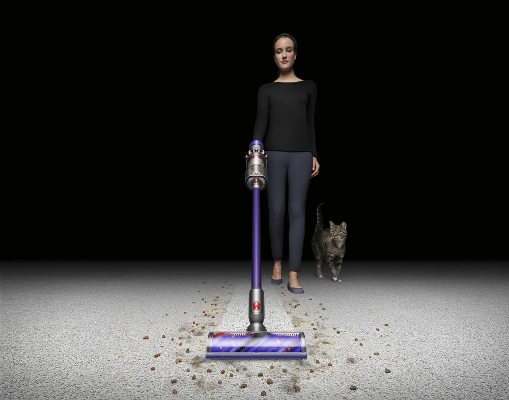 Mujer aspirando con una aspiradora Dyson. Gato y suelo sucio con escombros. Fondo negro.