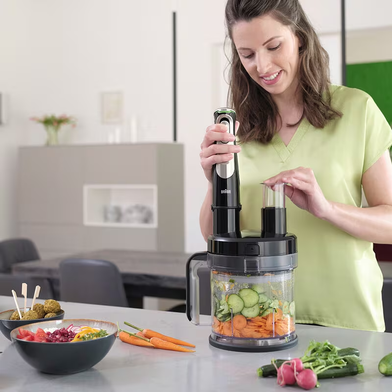 Une femme en chemise verte utilise un robot culinaire avec des légumes et de la nourriture.