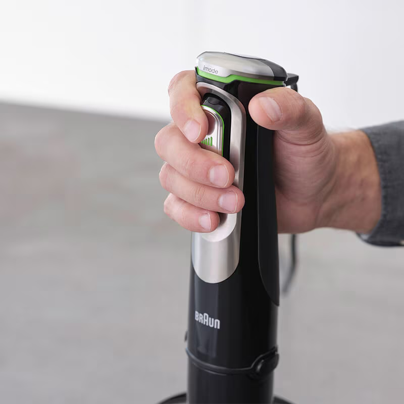 Main tenant un mixeur plongeant Braun noir et argent. Fond gris.