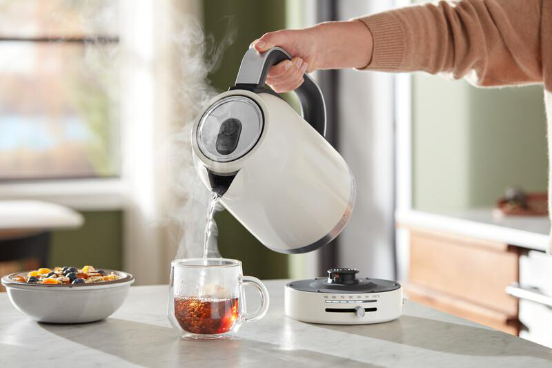 Person gießt Wasser aus einem weißen Wasserkocher in eine Glas-Tasse mit Tee.