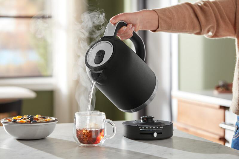 Eine Person gießt heißes Wasser aus einem schwarzen Wasserkocher in eine klare Tasse mit Tee. Eine Schüssel mit Essen steht daneben.