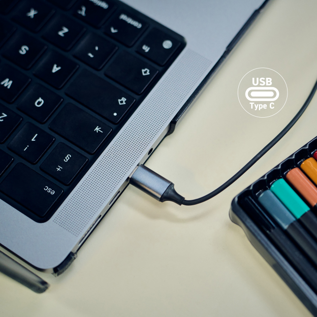 Egy laptop USB-C kábellel csatlakoztatva, színes ceruzák egy készlete a közelben.