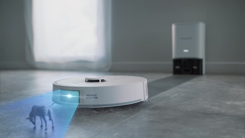 Fehér robotporszívó kék fénnyel, töltőállomás mellett, szürke csempézett padlón.