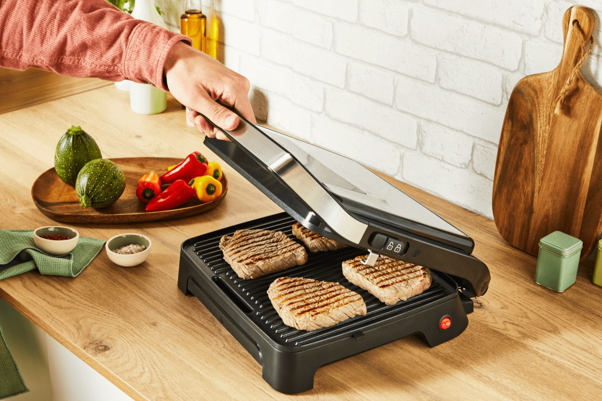 Grillezett steak a grillben, egy kéz nyitja. Zöldségek fa tálon a háttérben.