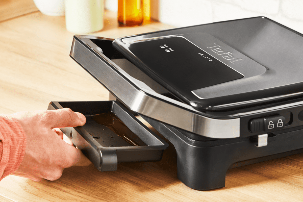 Egy személy kiveszi a zsírtálcát egy fekete-ezüst Tefal grillből. A tálca tele van zsírral.