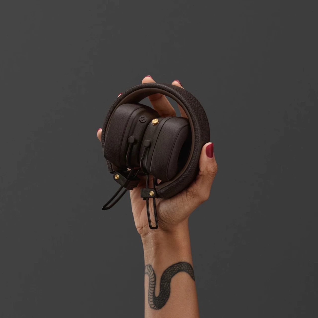 Dłoń trzyma złożone brązowe słuchawki ze złotymi akcentami na ciemnoszarym tle.