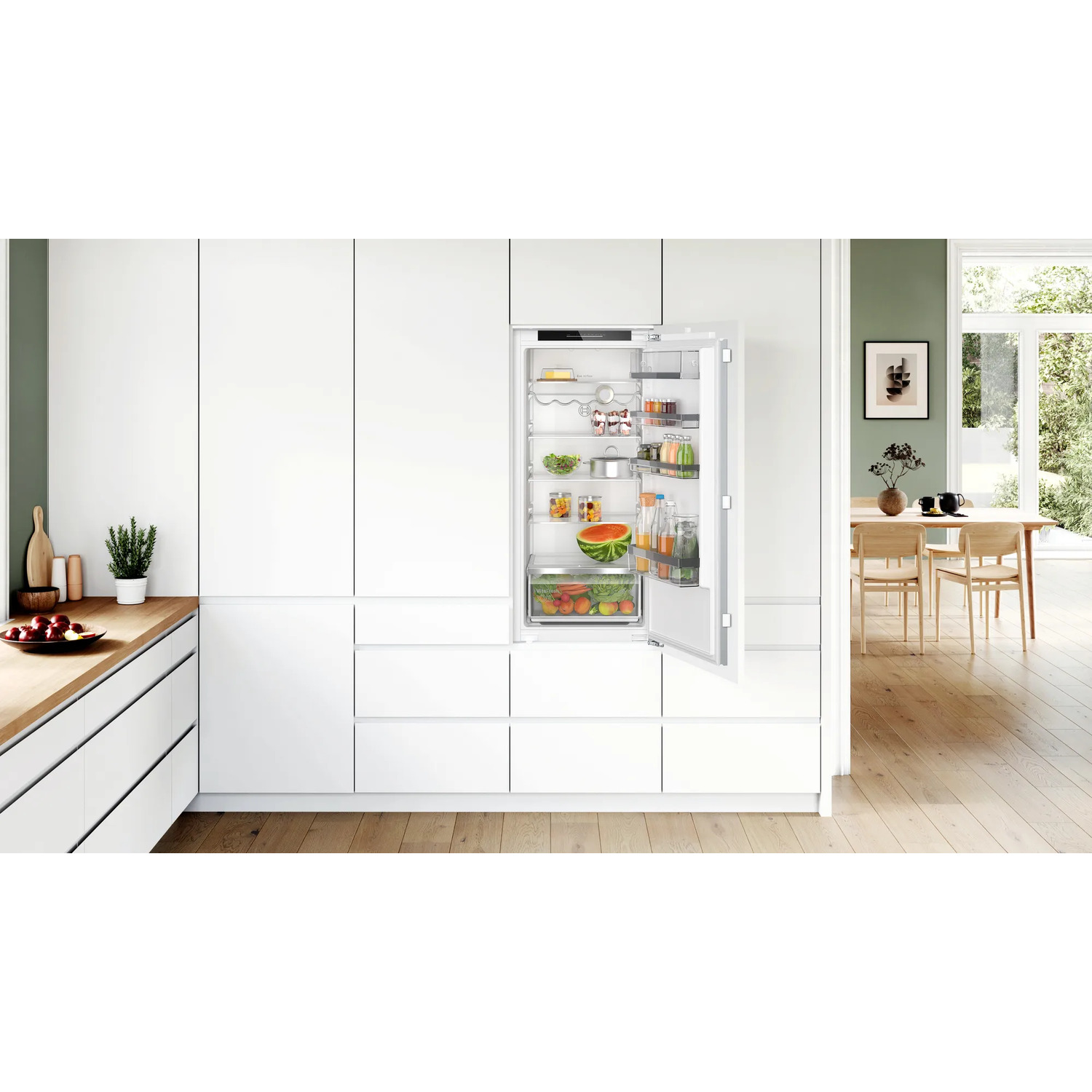 Modern white kitchen. Refrigerator door open, revealing shelves with food. Wooden countertop with plants.