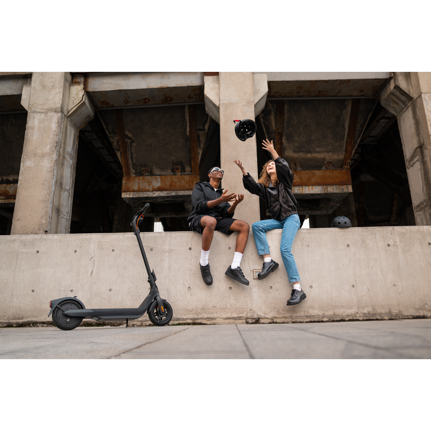 Deux personnes sur une corniche en béton, l'une lançant un casque en l'air, près d'un scooter et d'un bâtiment.