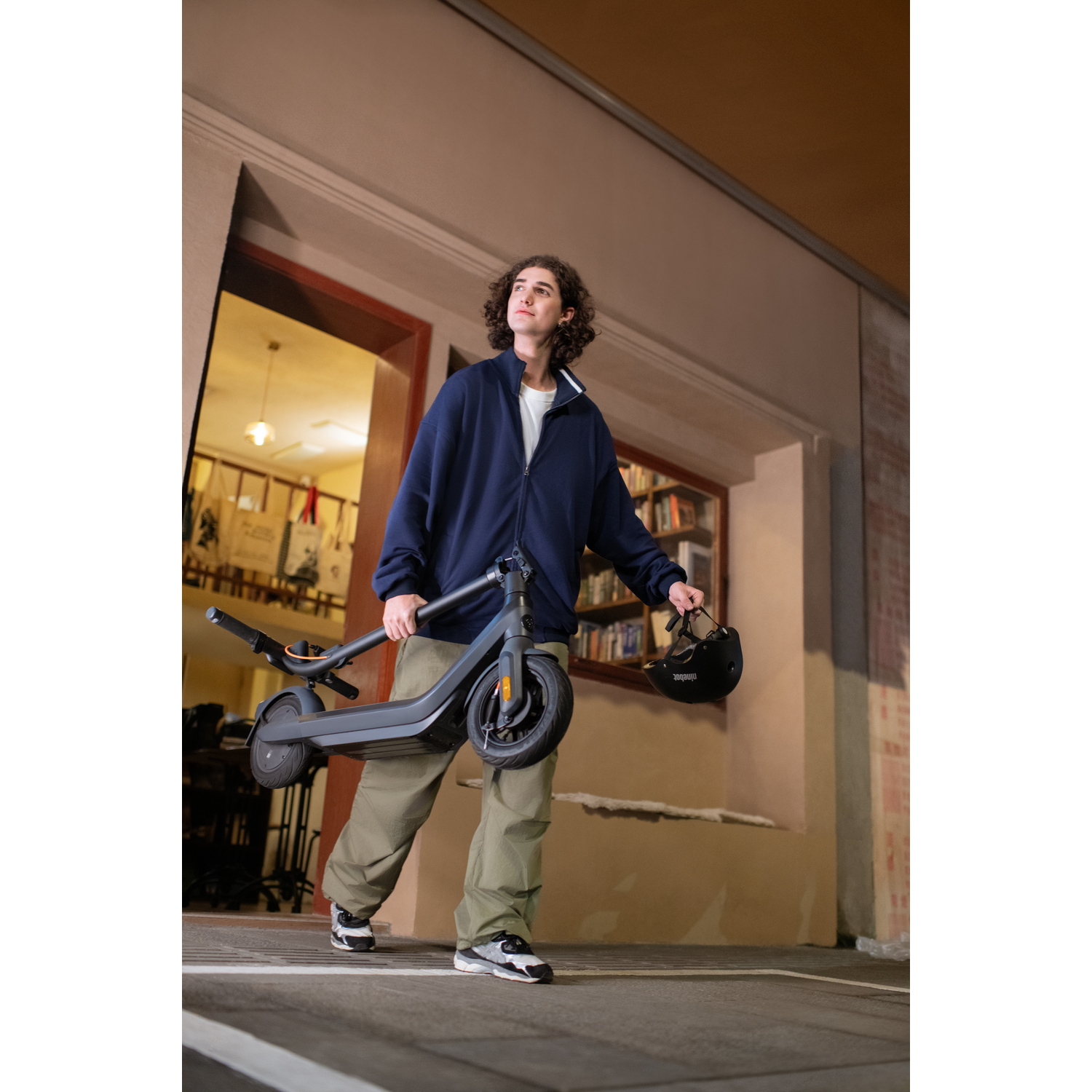 Un jeune homme porte un scooter noir plié. Il marche dans une rue la nuit.