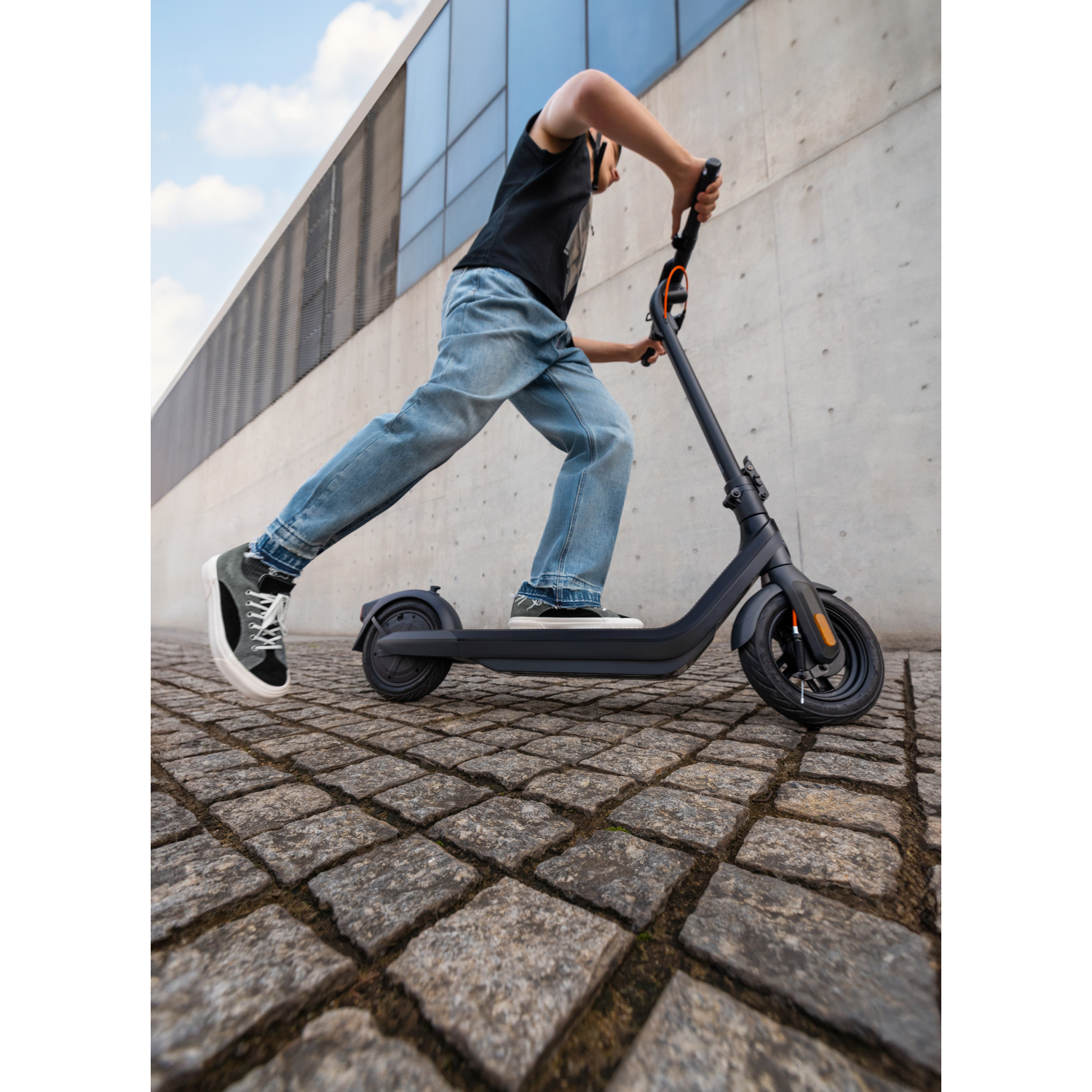 Personne conduisant une trottinette noire sur une rue pavée. Bâtiment gris en arrière-plan.