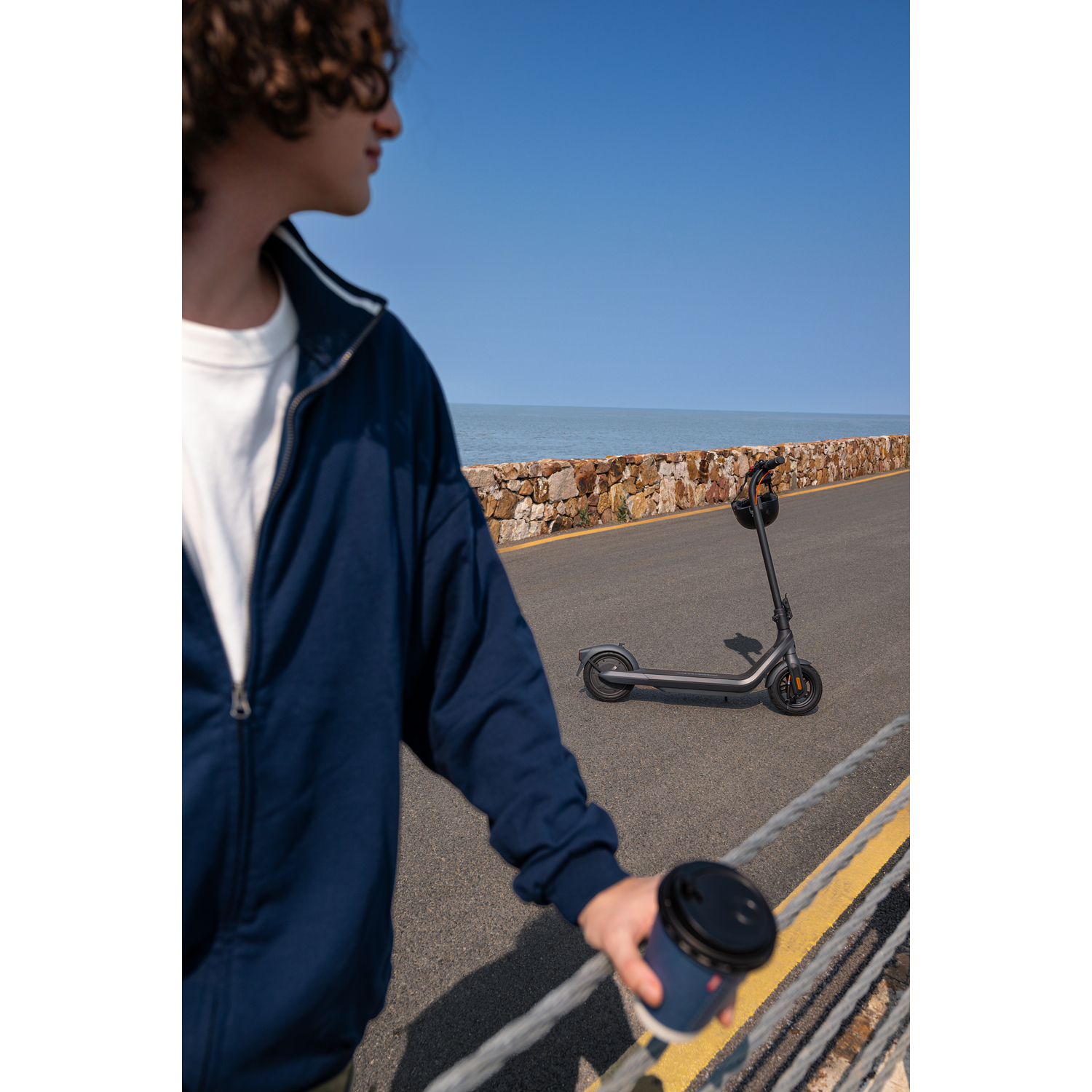 Une personne avec des lunettes de soleil et une veste bleue se tient près d'un scooter et tient une tasse de café, avec la mer et le ciel en arrière-plan.