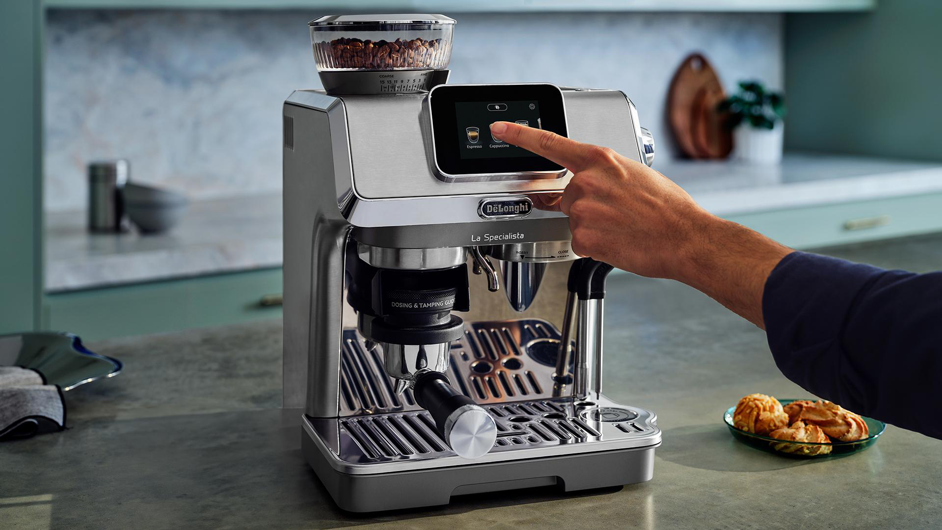 Eine Person berührt den Bildschirm einer Kaffeemaschine. Oben ist eine Kaffeemühle.