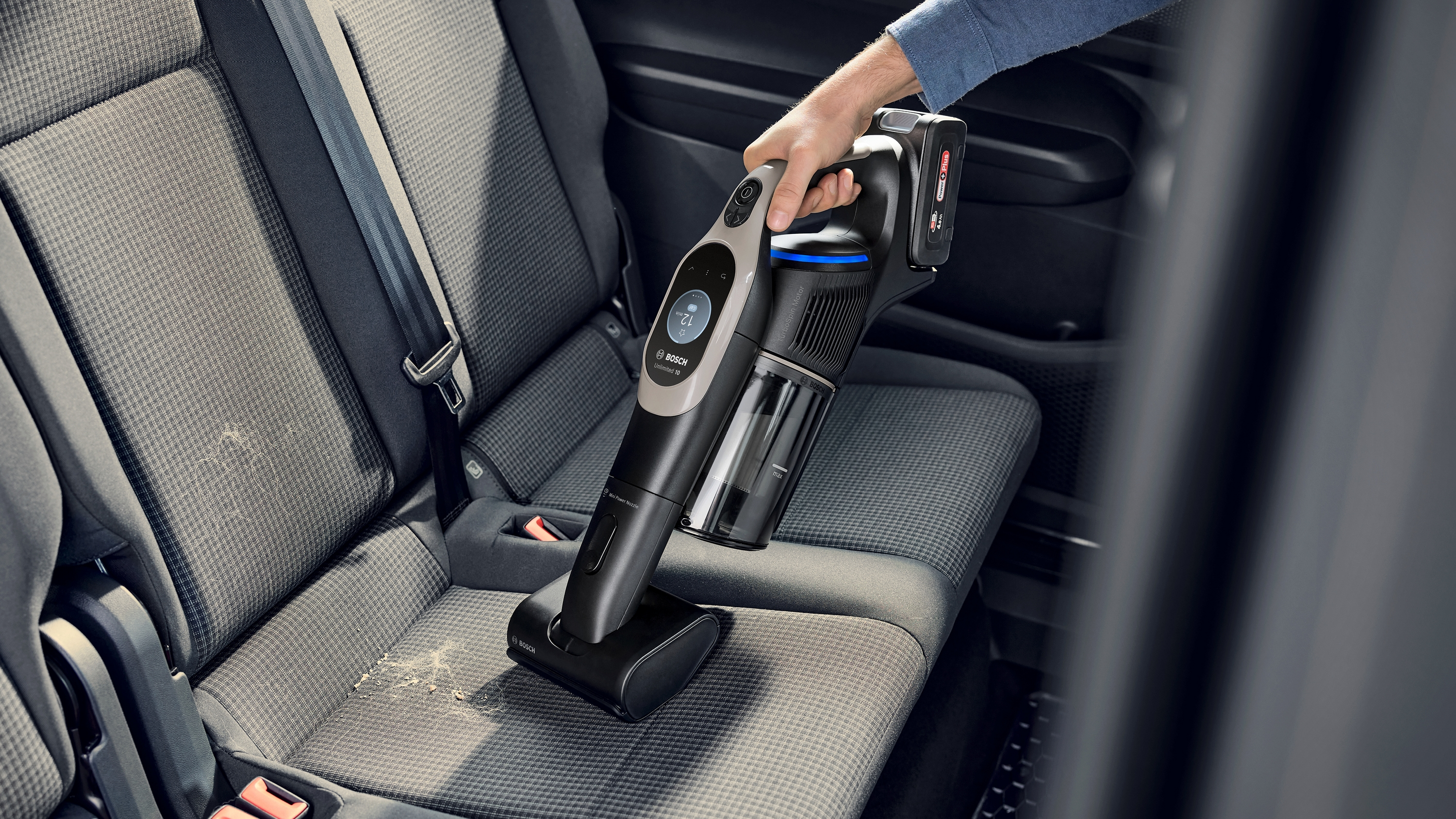 Eine Person, die mit einem Handstaubsauger einen Autositz absaugt. Der Sitz ist grau.