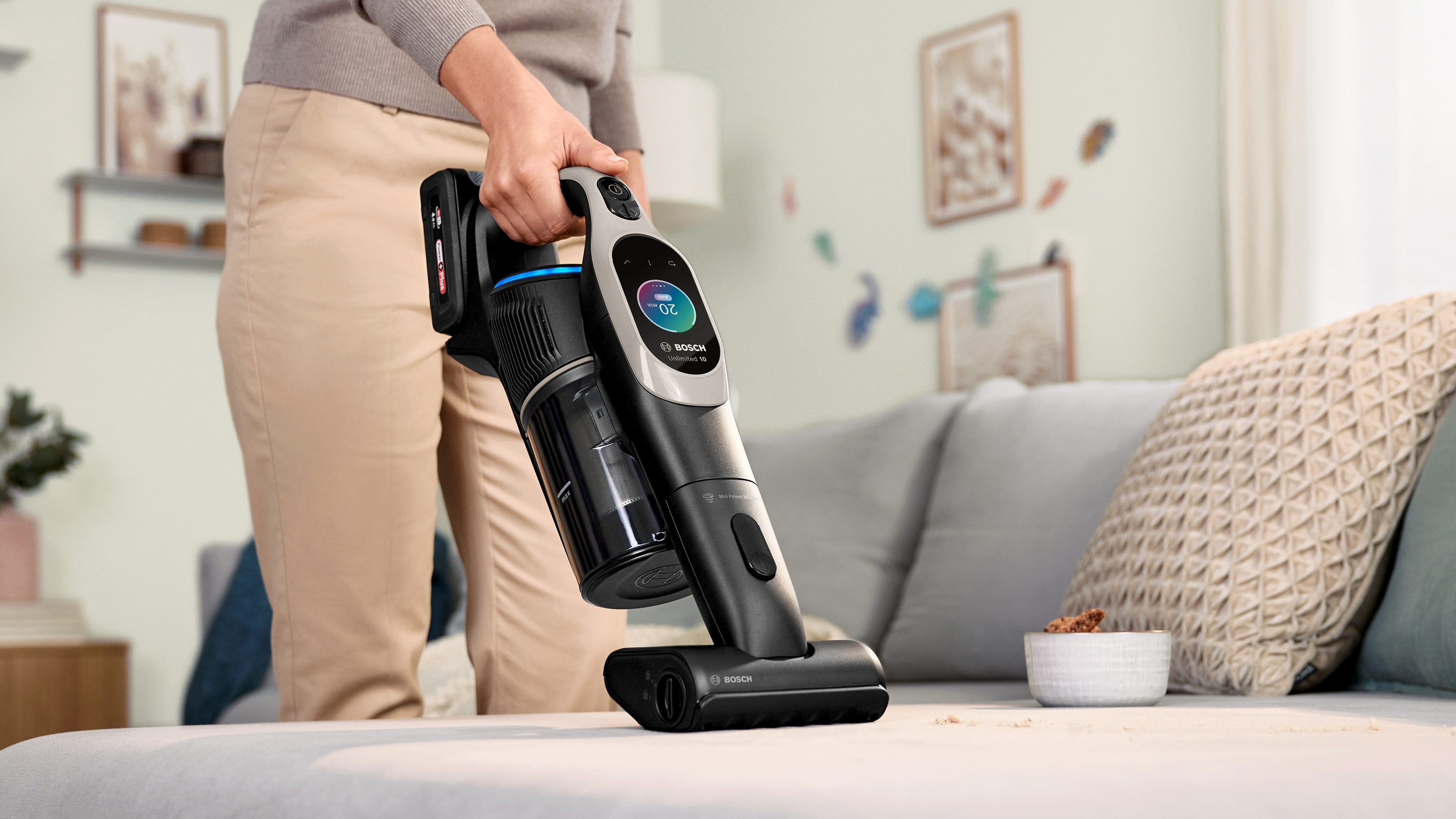 Eine Frau hält einen schwarz-grauen Staubsauger auf einem Sofa.