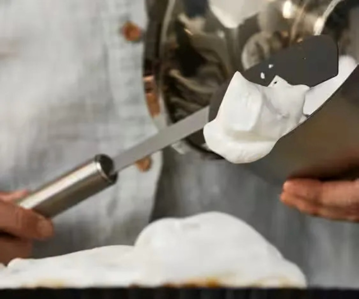 Szpatułka nabiera bezy z miski i przenosi ją na ciemną blachę do pieczenia.