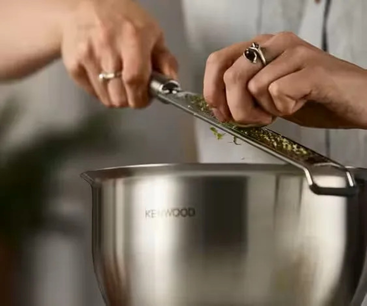 Dłonie ścierają zioła do miski ze stali nierdzewnej za pomocą tarki. Misa ma etykietę.