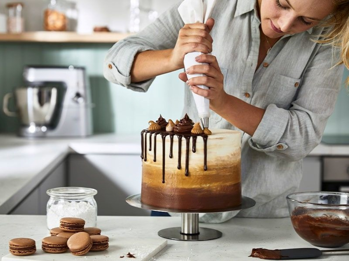 Kobieta dekoruje warstwowe ciasto polewą czekoladową i lukrem. Makaroniki i miska do mieszania znajdują się na pierwszym planie.