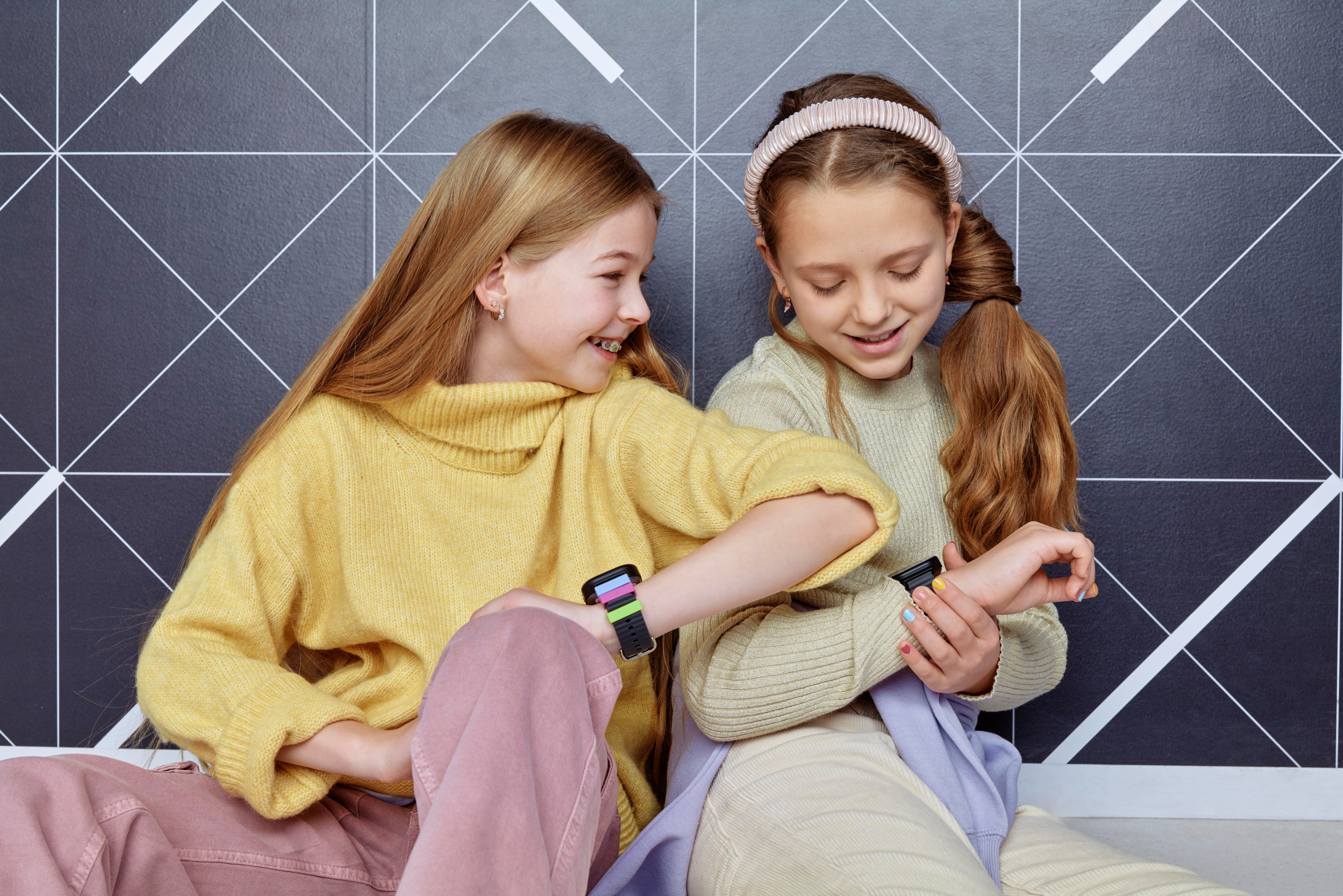 Zwei junge Mädchen schauen sich Smartwatches an. Eines trägt einen gelben Pullover, das andere ein rosa Stirnband.