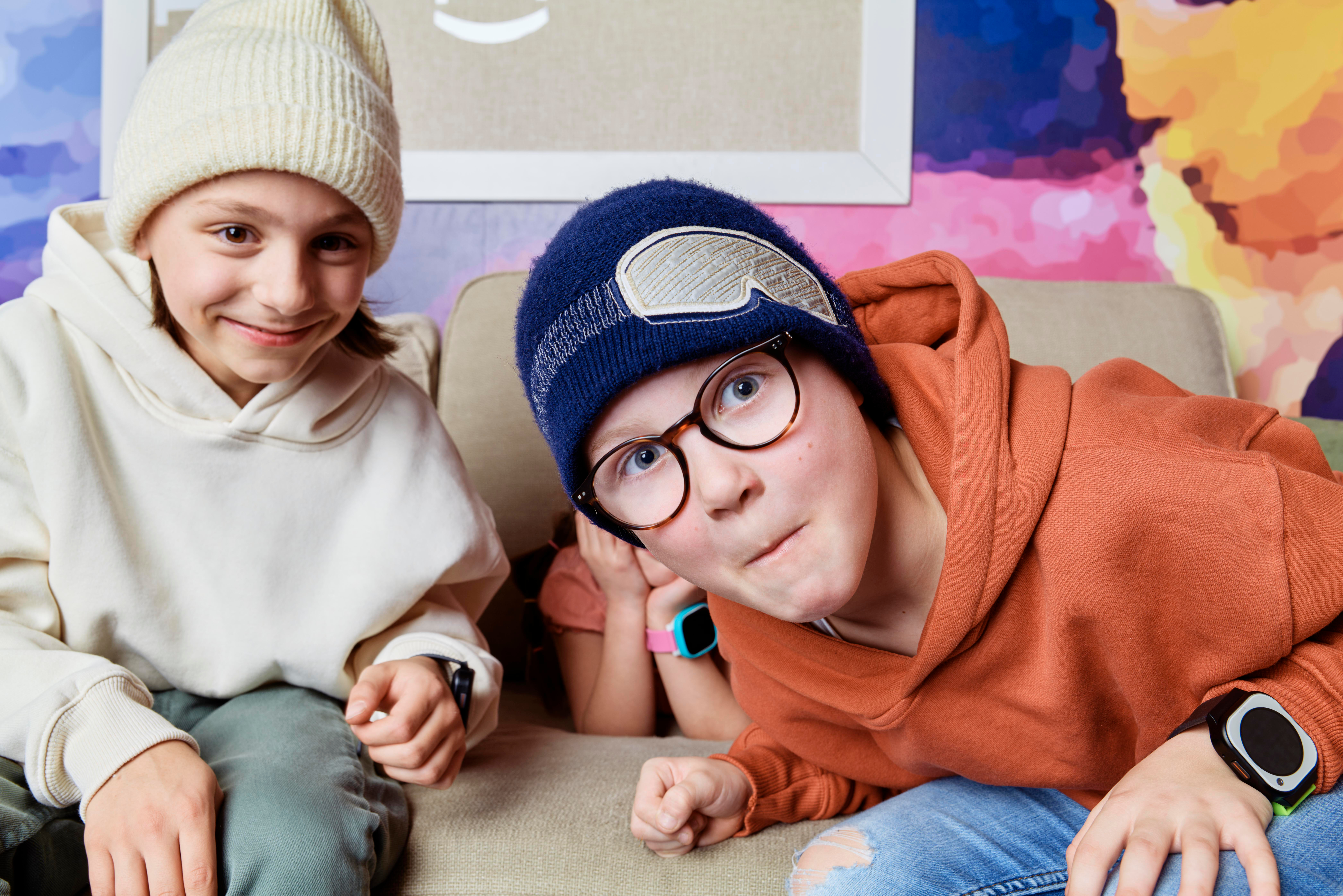 Zwei lächelnde Kinder, Wintermützen, sitzen auf Couch.