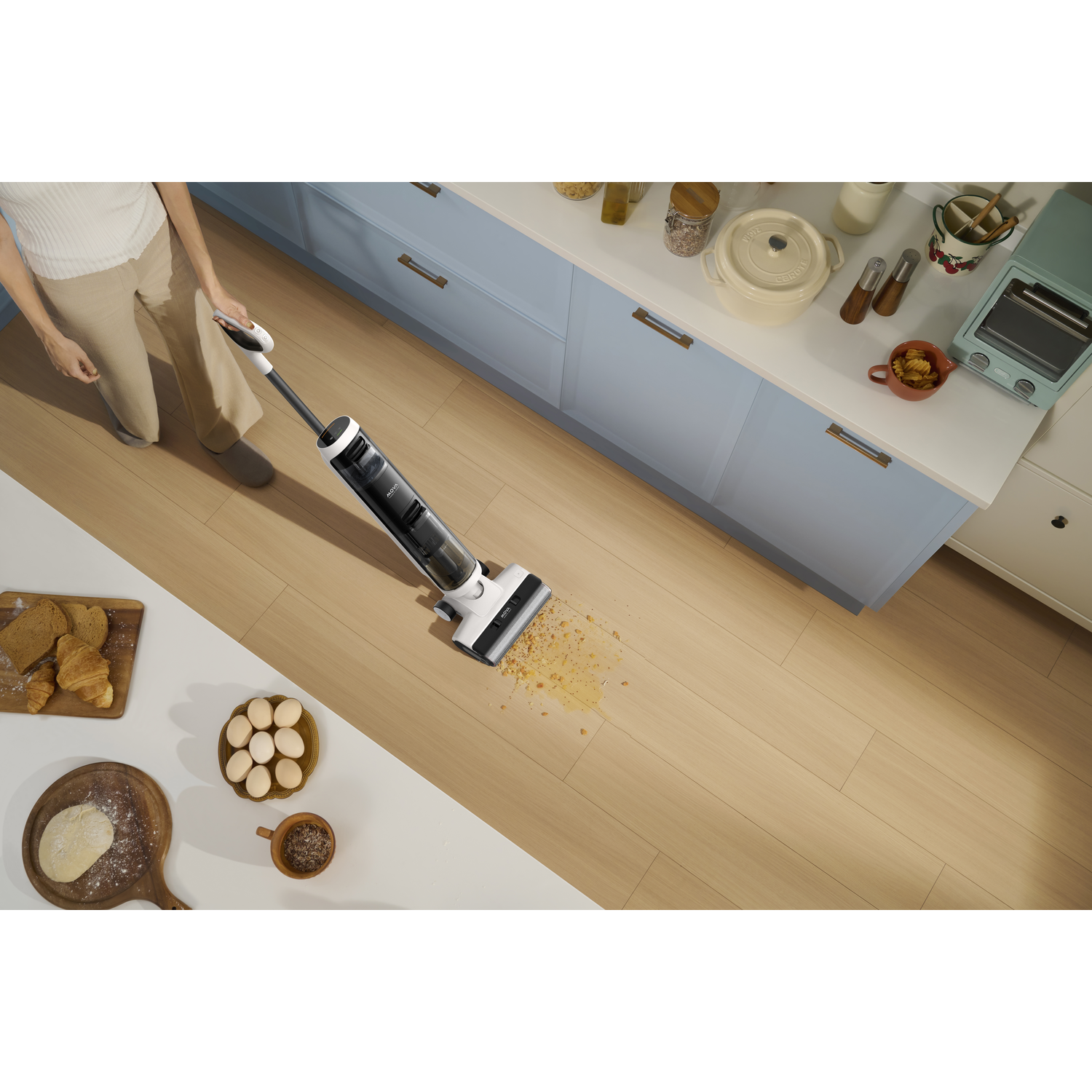 Person vacuuming a kitchen floor with a white and black vacuum, food and eggs on the counter.