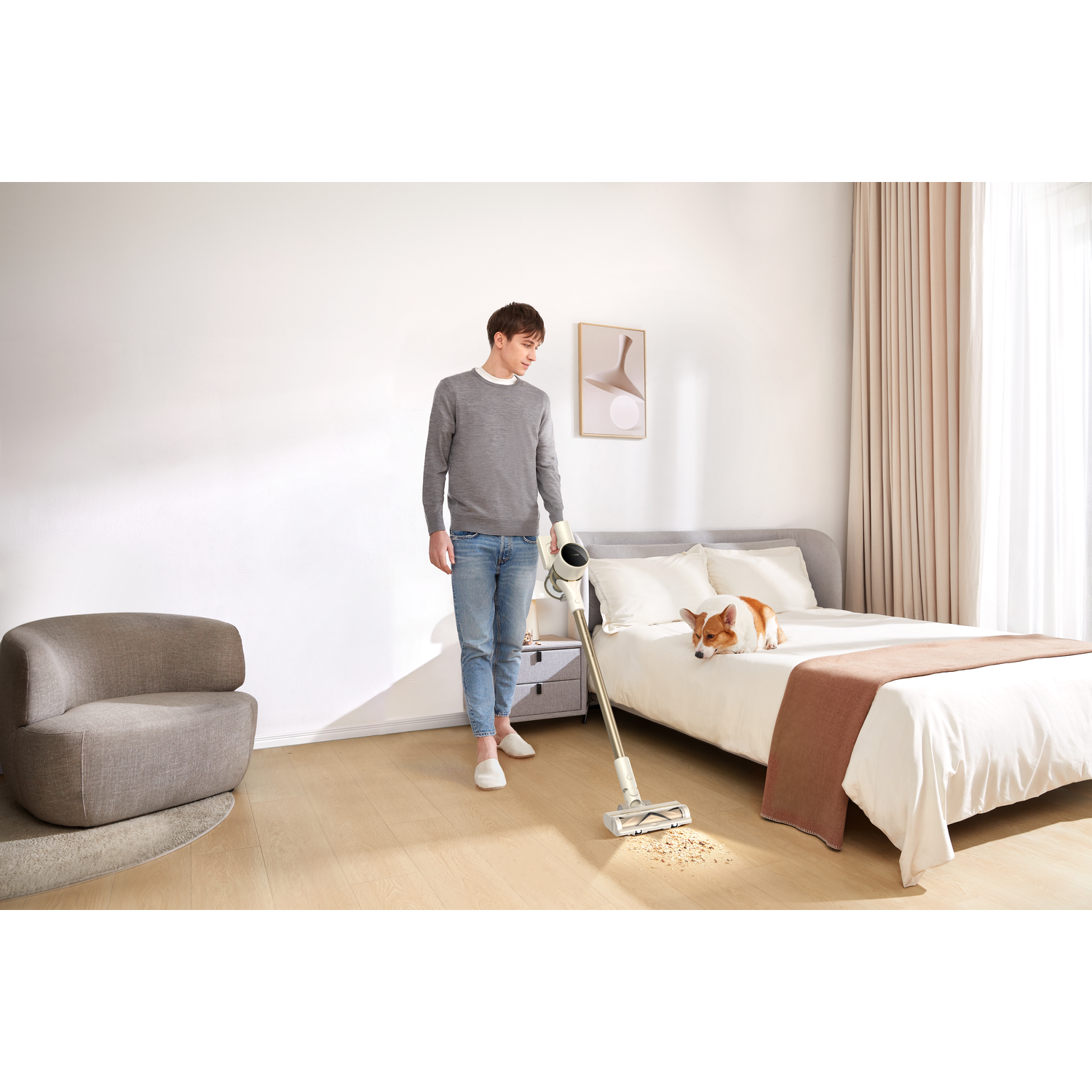 Man vacuuming a wooden floor with a cordless vacuum; dog on bed in the background.