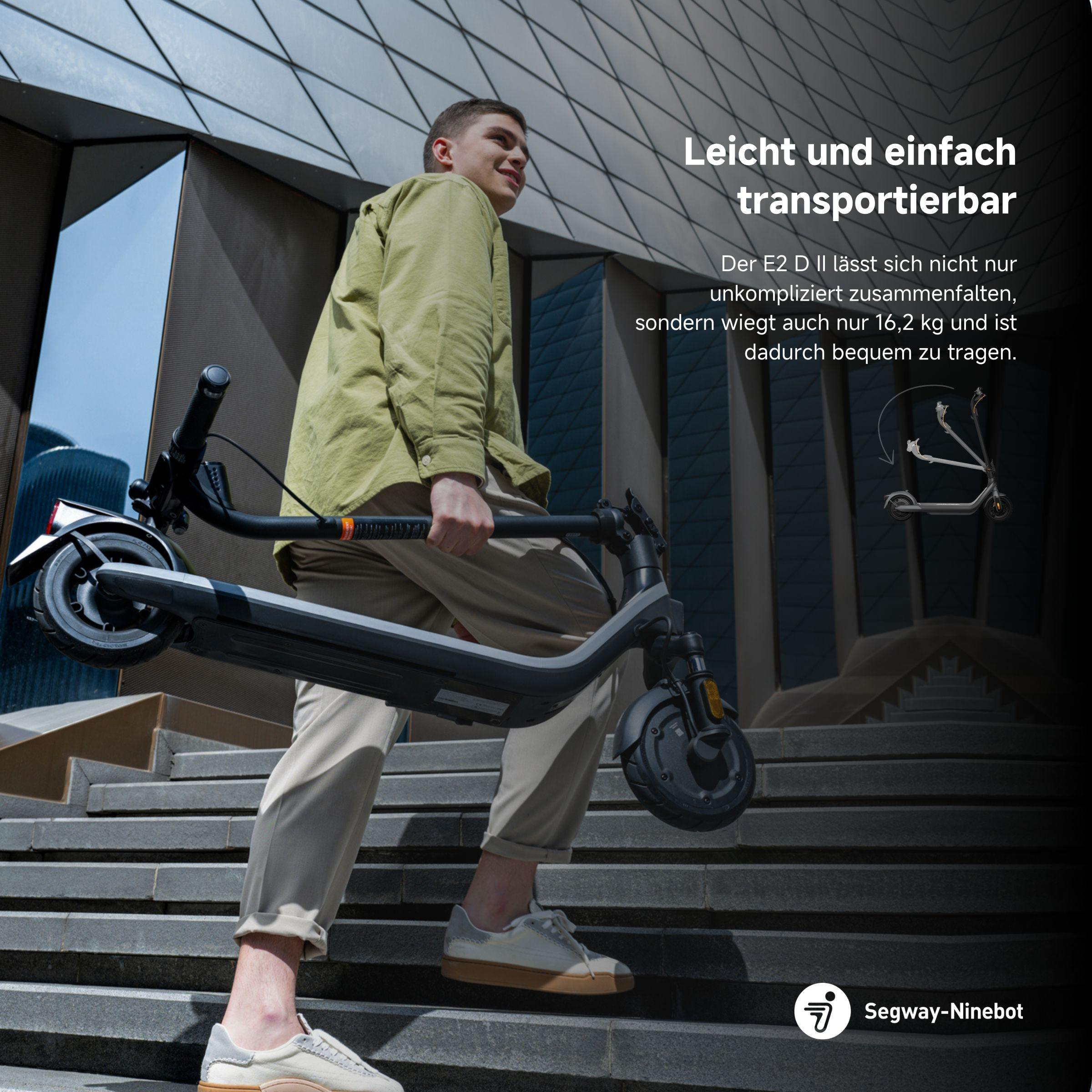 Mann trägt einen gefalteten schwarzen Roller die Treppe hoch und lächelt. Hintergrund: modernes Gebäude.