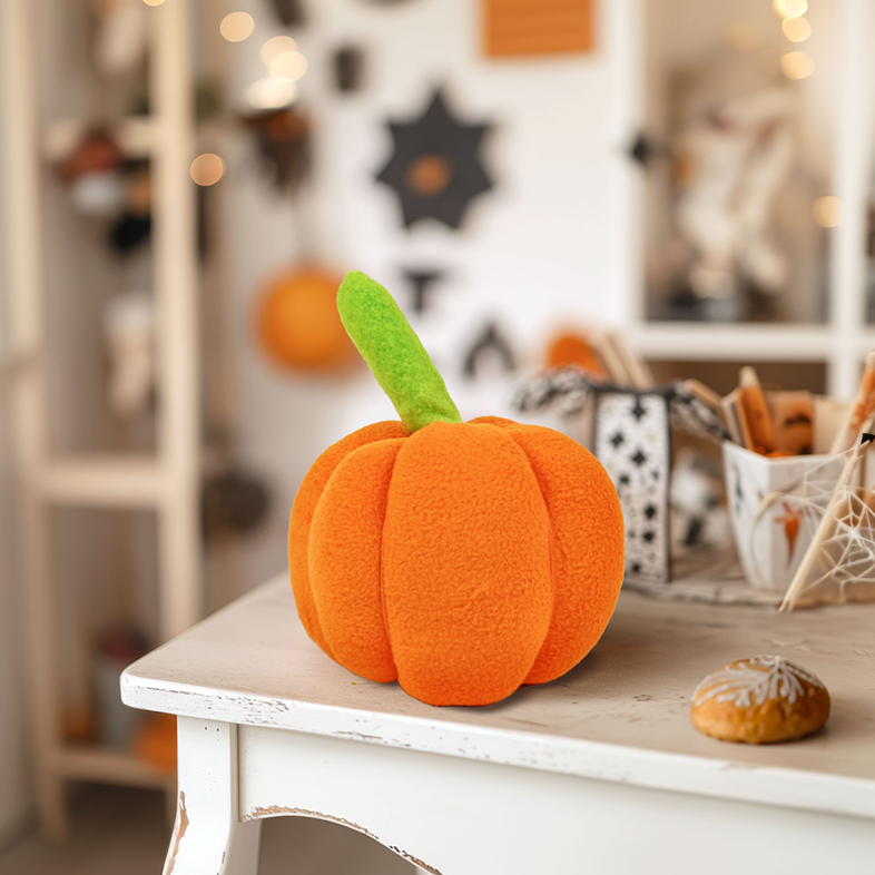 Egy plüss narancssárga tök zöld szárral. Egy fehér asztalon van. Háttér Halloween dekorációval.