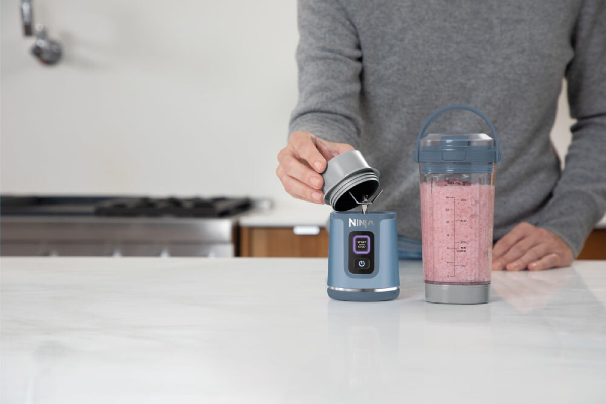 Osoba dodaje składniki do niebieskiego blendera, obok pojemnika z koktajlem.