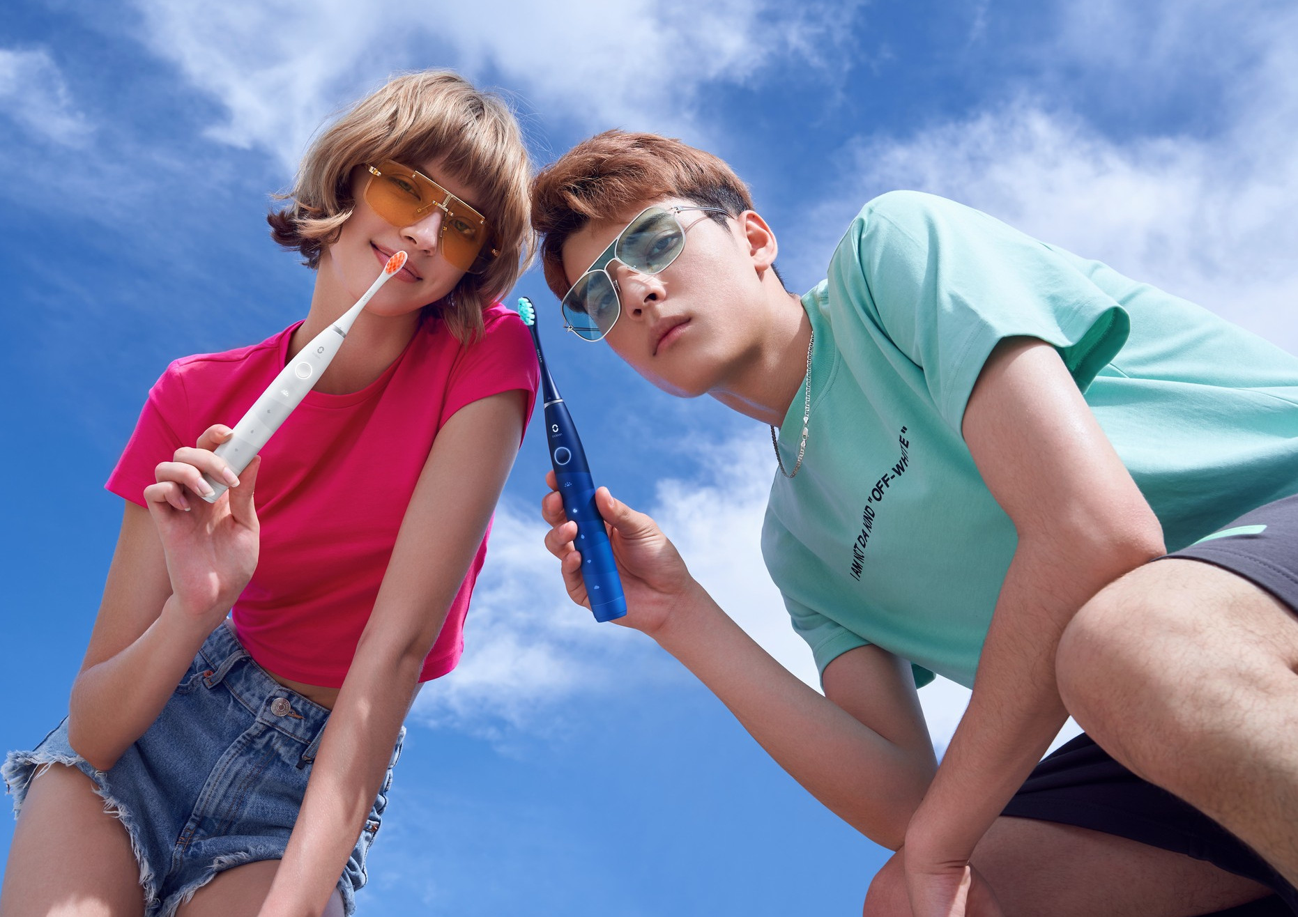 Dwie osoby trzymają elektryczne szczoteczki do zębów, uśmiechają się. Błękitne niebo w tle.