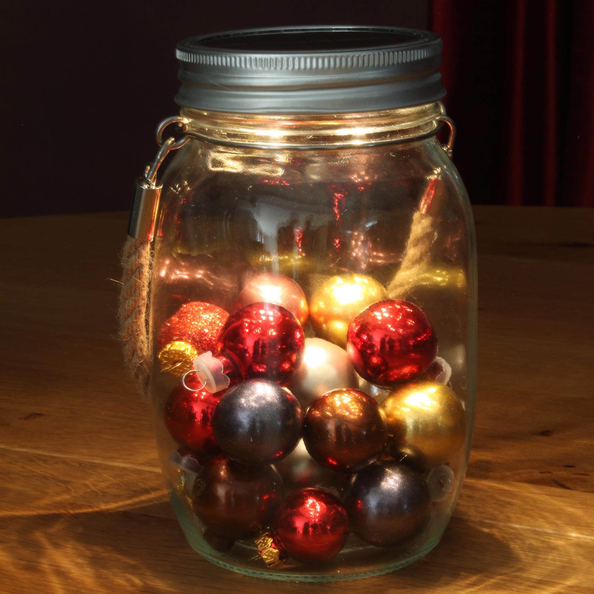 Ein Glas mit roten, silbernen und goldenen Weihnachtskugeln, von innen beleuchtet, auf Holz.
