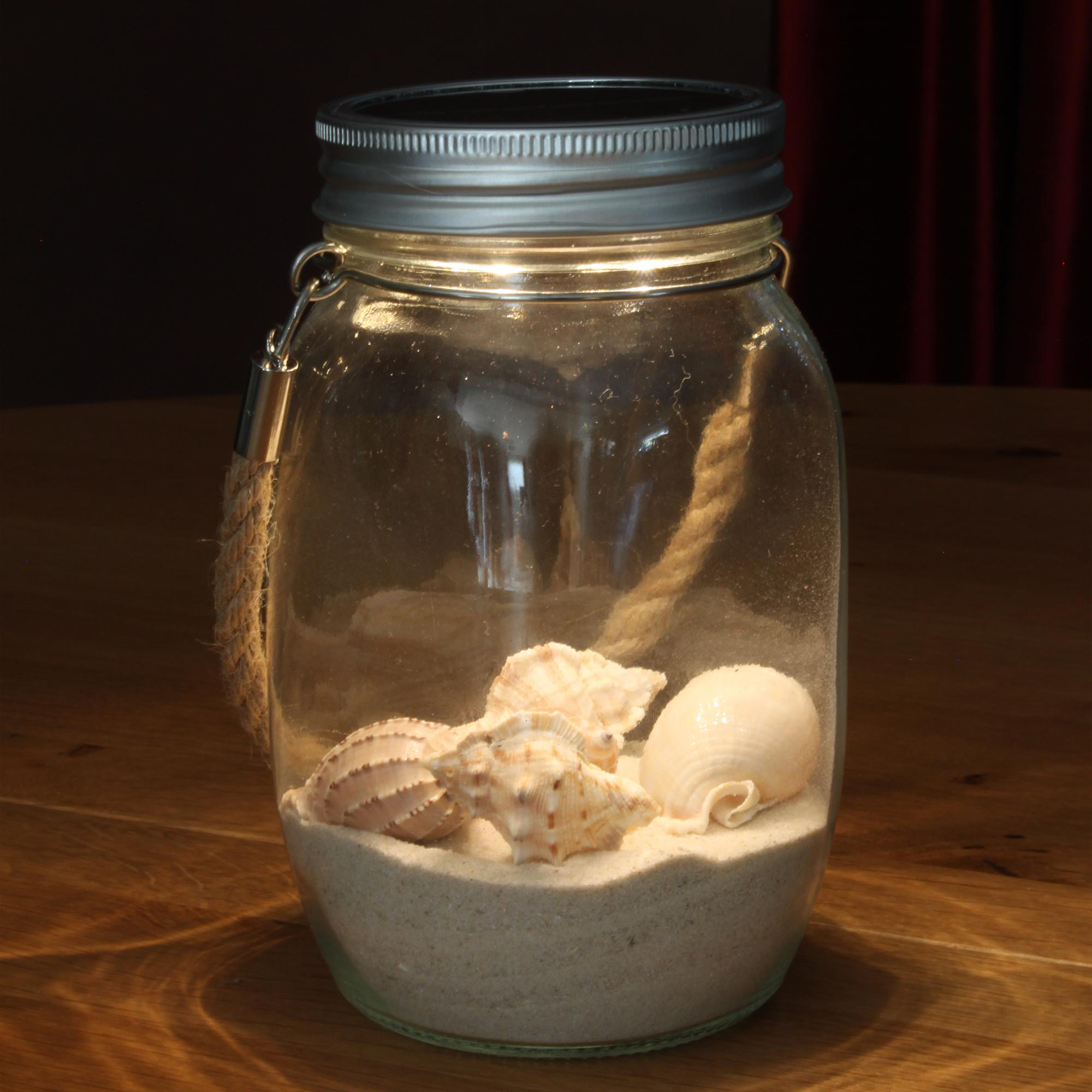 Ein Glas mit Sand und Muscheln, von innen beleuchtet, auf Holz, mit dunklem Hintergrund.