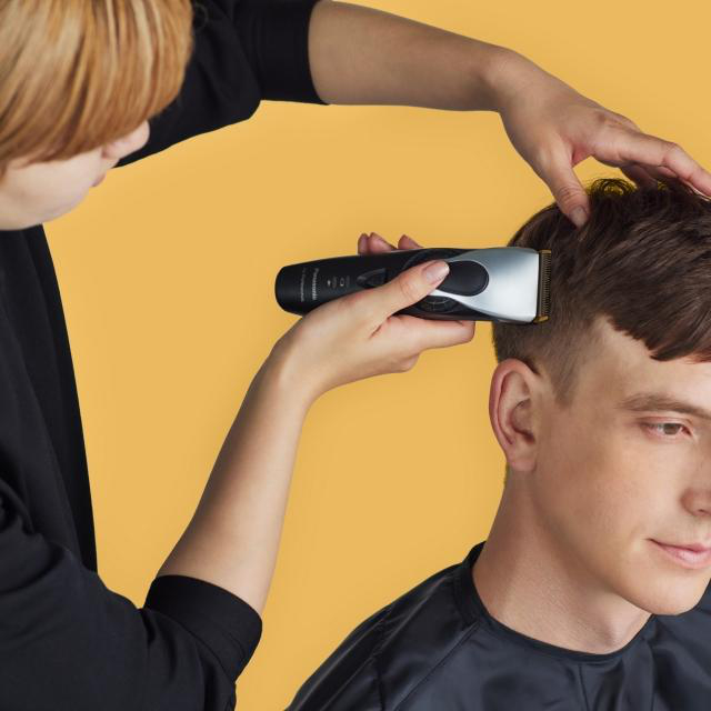 Ein Friseur schneidet einem Mann die Haare mit einem Haarschneider, gelber Hintergrund.
