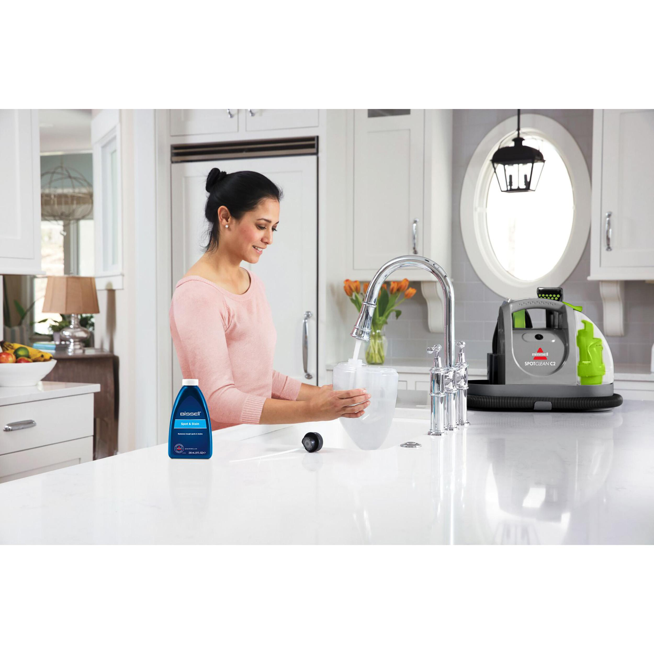 Eine Frau füllt einen Wassertank an einem Küchenbecken, mit einer Bissell SpotClean Maschine und Reinigerflasche.