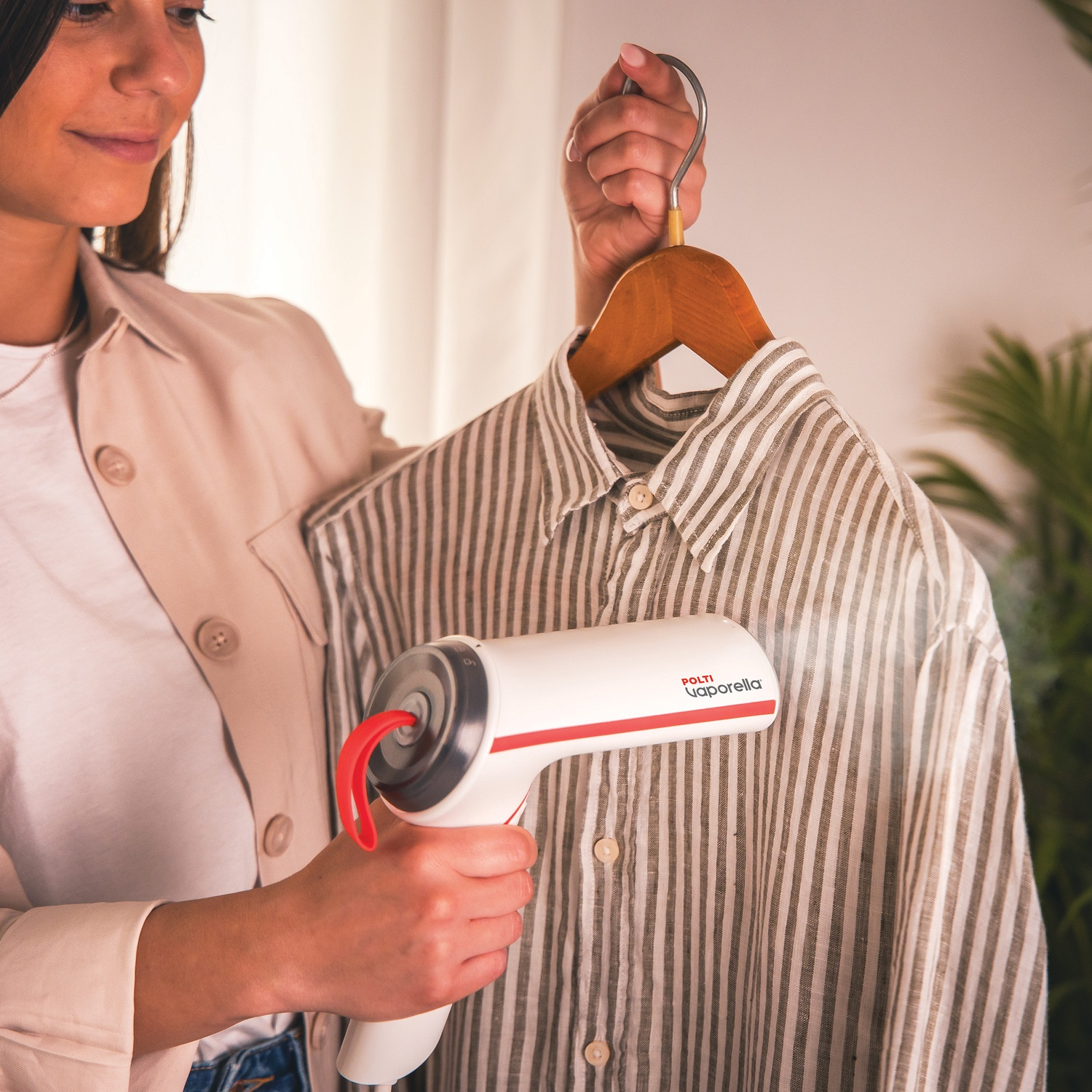 Una donna usa un vaporizzatore bianco e rosso per stirare una camicia a righe su una gruccia di legno.
