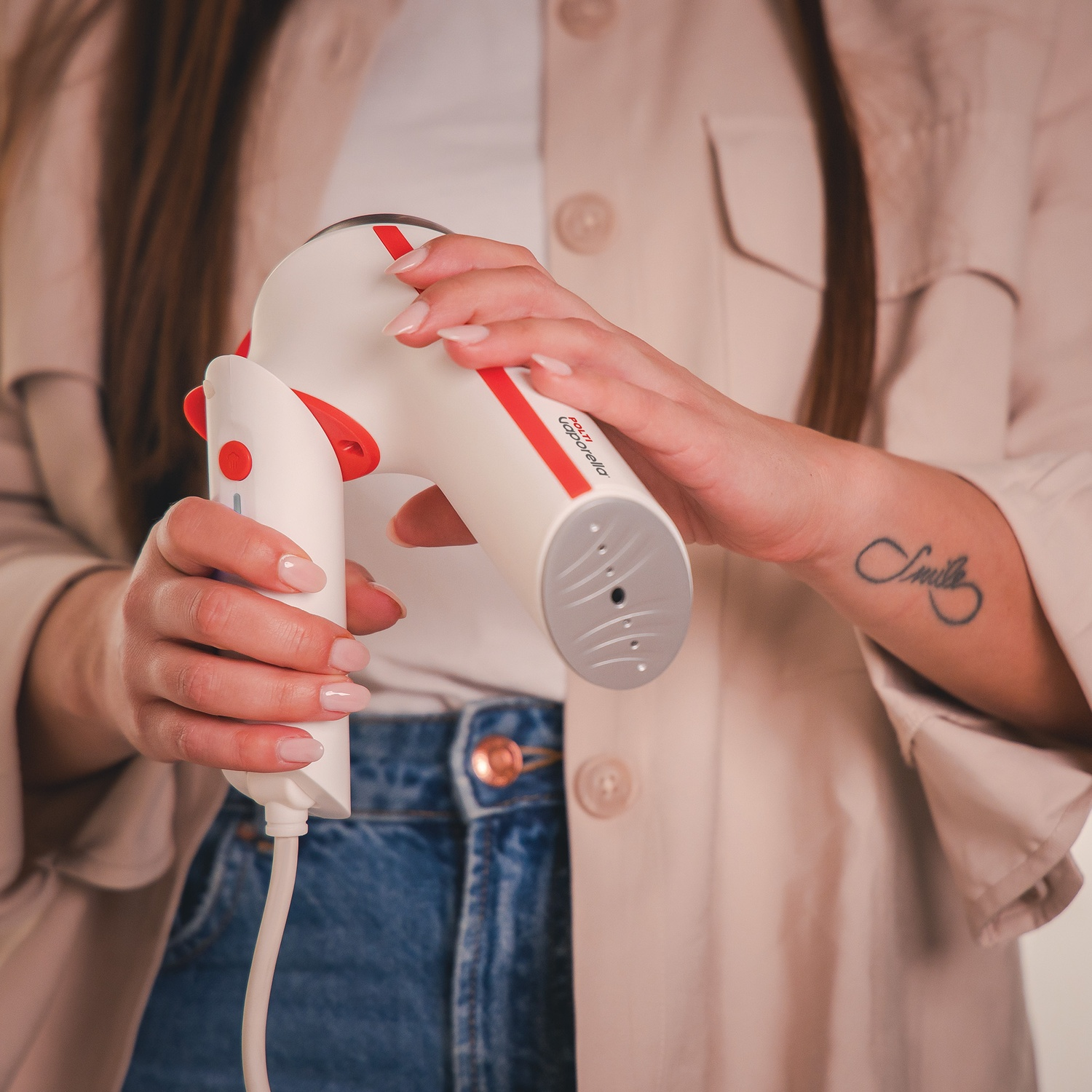 Persona che tiene un vaporizzatore per indumenti bianco e rosso, tatuaggio visibile, davanti a giacca beige.