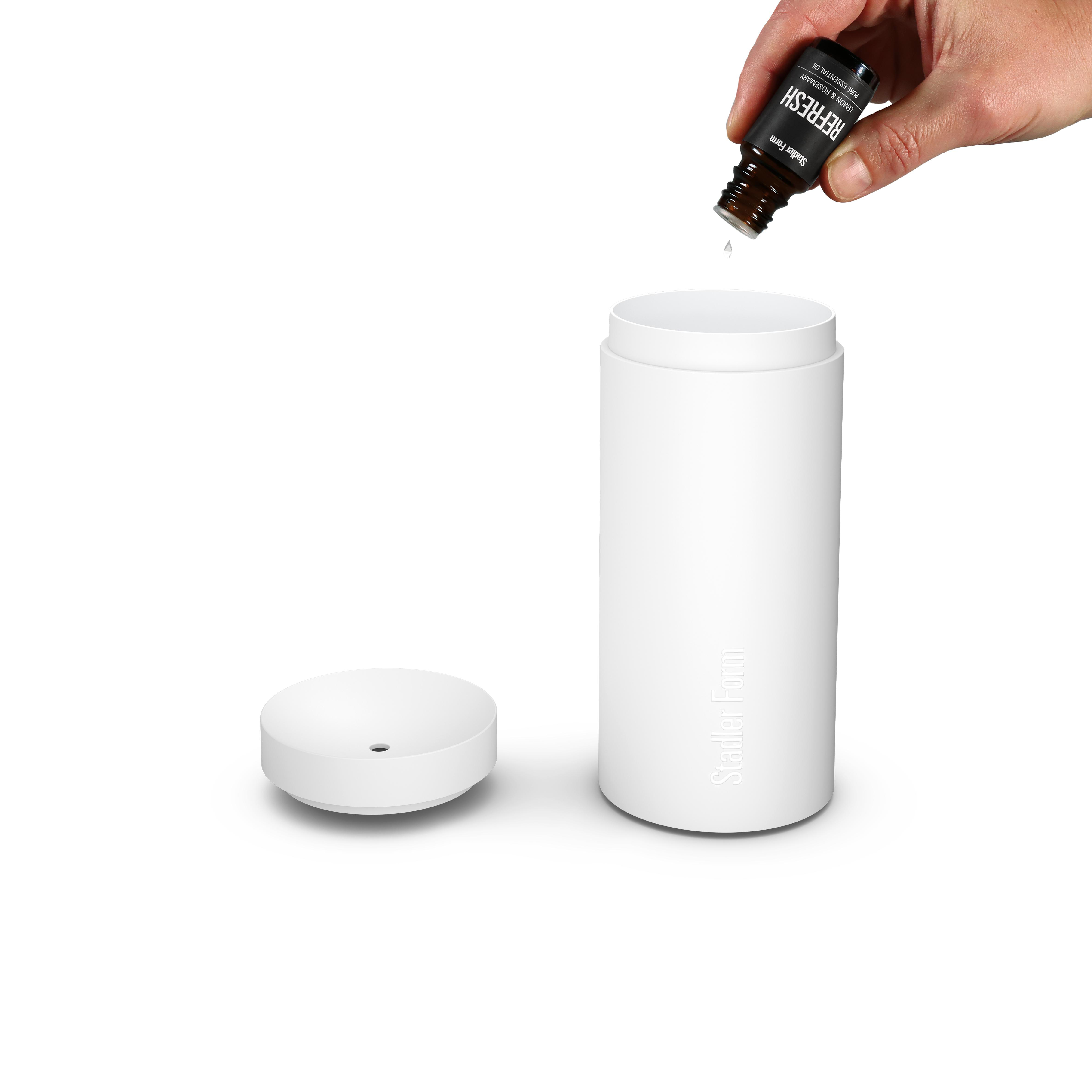 Hand gießt Öl aus einer Flasche in einen weißen Diffusor, mit separatem Deckel daneben.