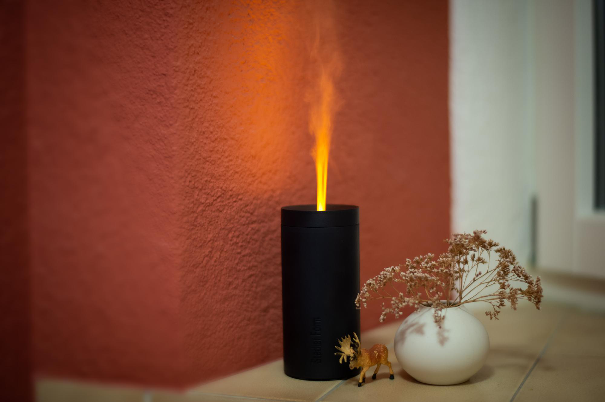 Schwarzer Diffusor mit orangenem, flammenartigen Dampf. Weiße Vase mit Trockenblumen und ein kleines Reh.