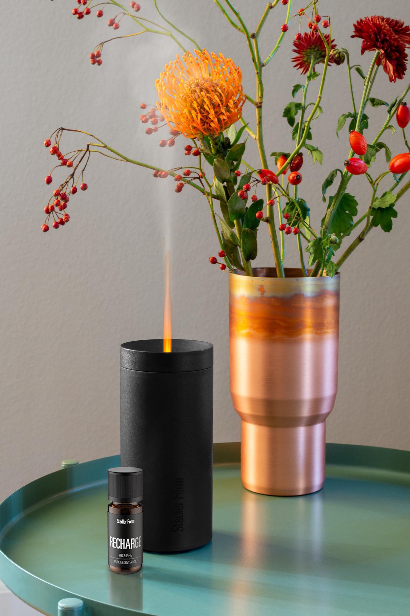 Schwarzer Diffusor mit Ölflasche und Kupfervase mit Blumen auf grünem Tisch.