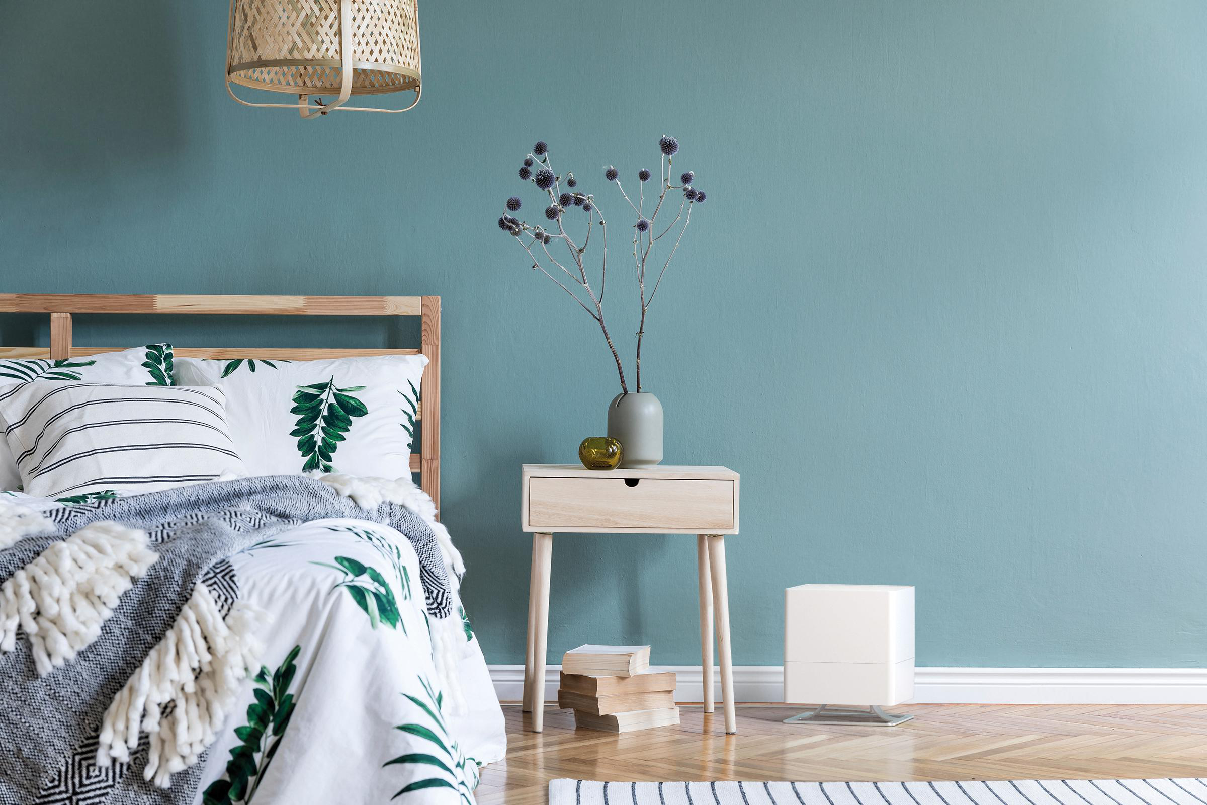 Schlafzimmer. Bett mit Blumendruck, Nachttisch mit Vase und blaue Wand im Hintergrund.