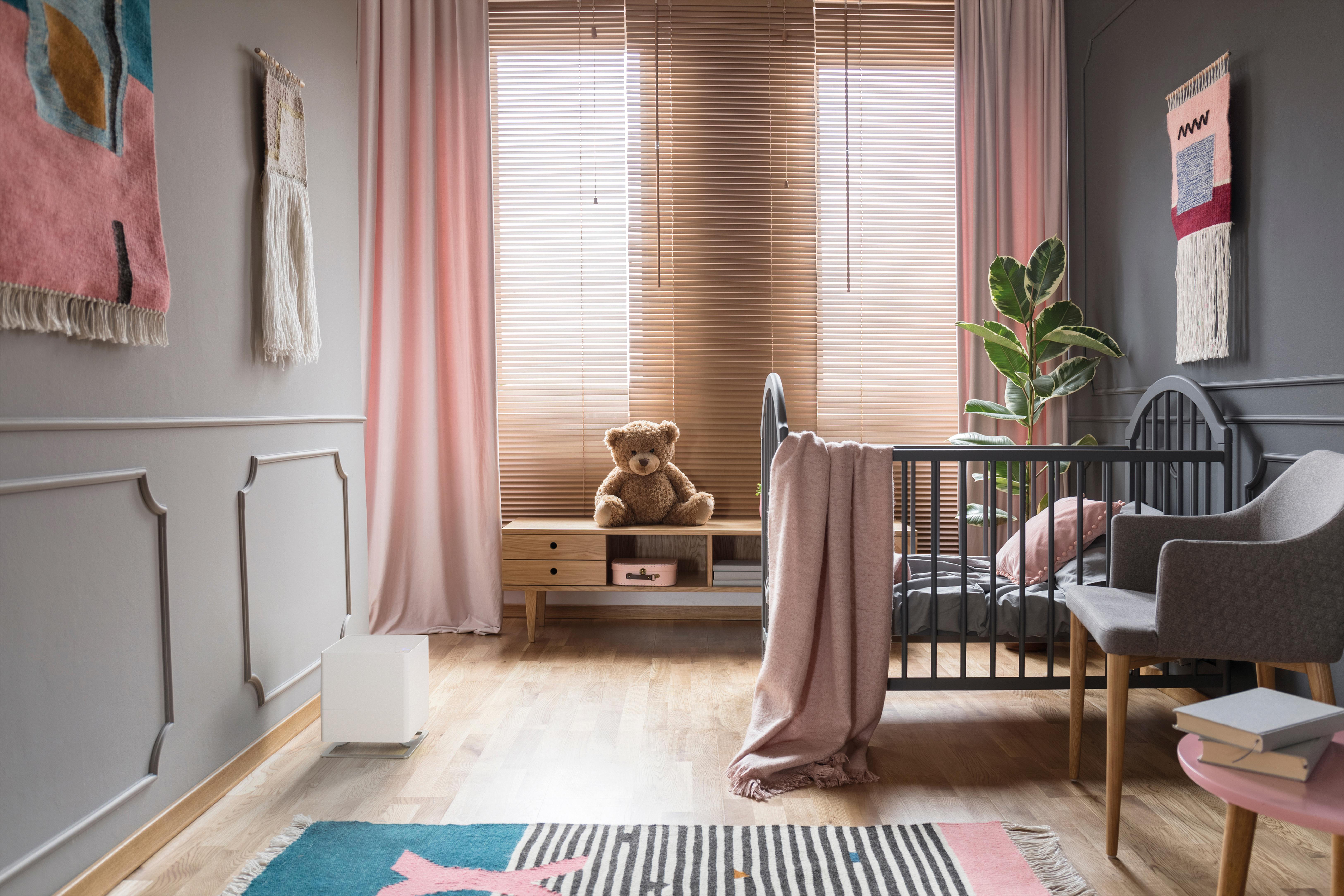 Ein Kinderzimmer mit grauen Wänden, rosa Vorhängen, einem Kinderbett, einem Teddybären und Wandbehängen.