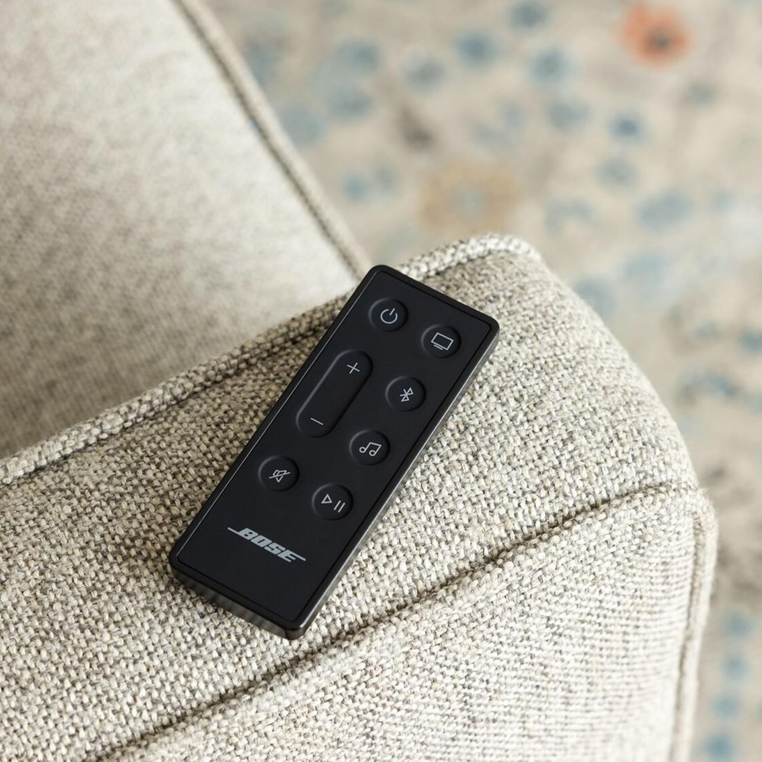 Un telecomando Bose nero su una poltrona grigia strutturata, con sfondo sfocato.