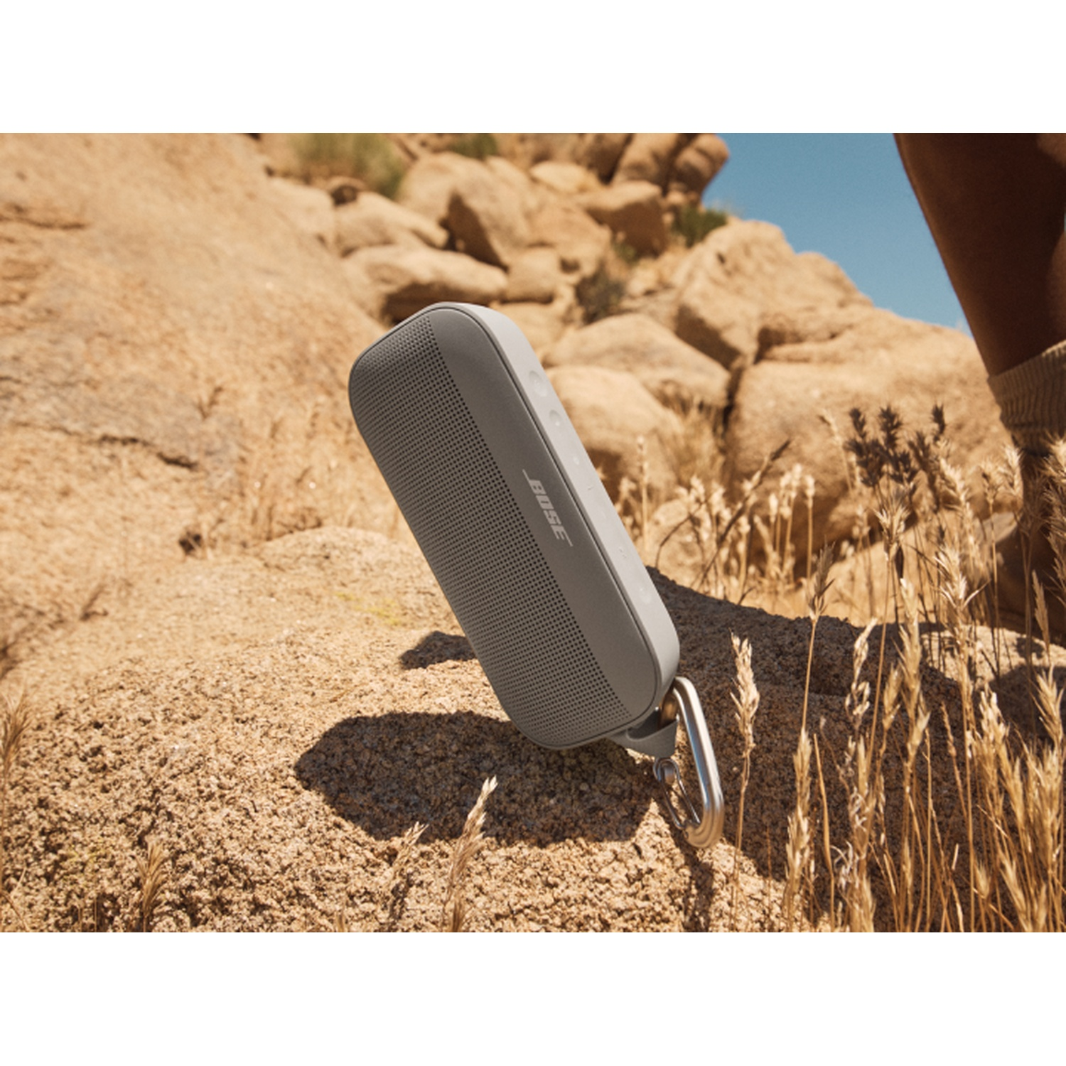 Altoparlante grigio con clip argentata su roccia beige. Sfondo: rocce e cielo blu.