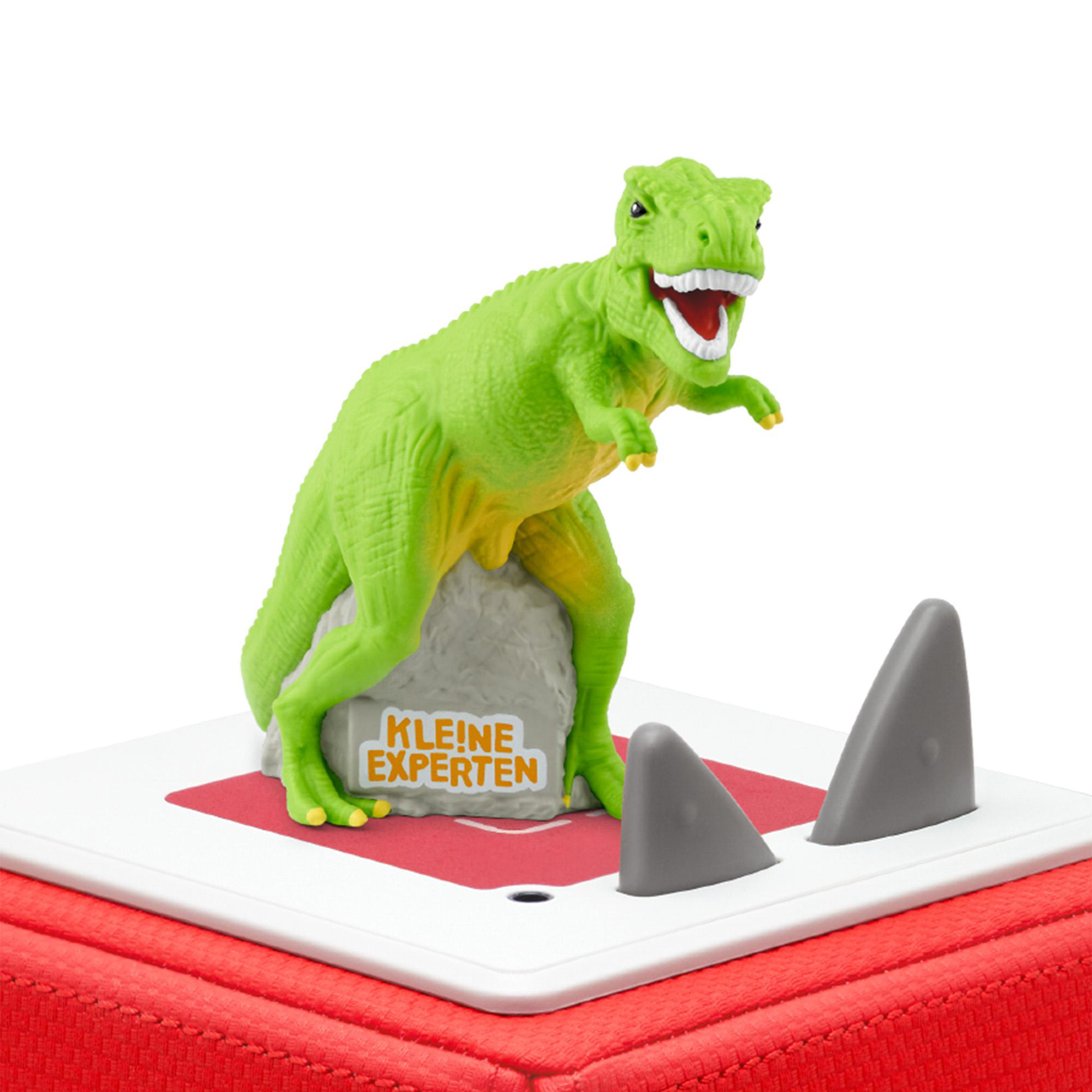 Ein grünes Dinosaurierspielzeug auf einer roten Plattform mit zwei grauen Haifischflossen. Das Schild lautet 'KLEINE EXPERTEN'.