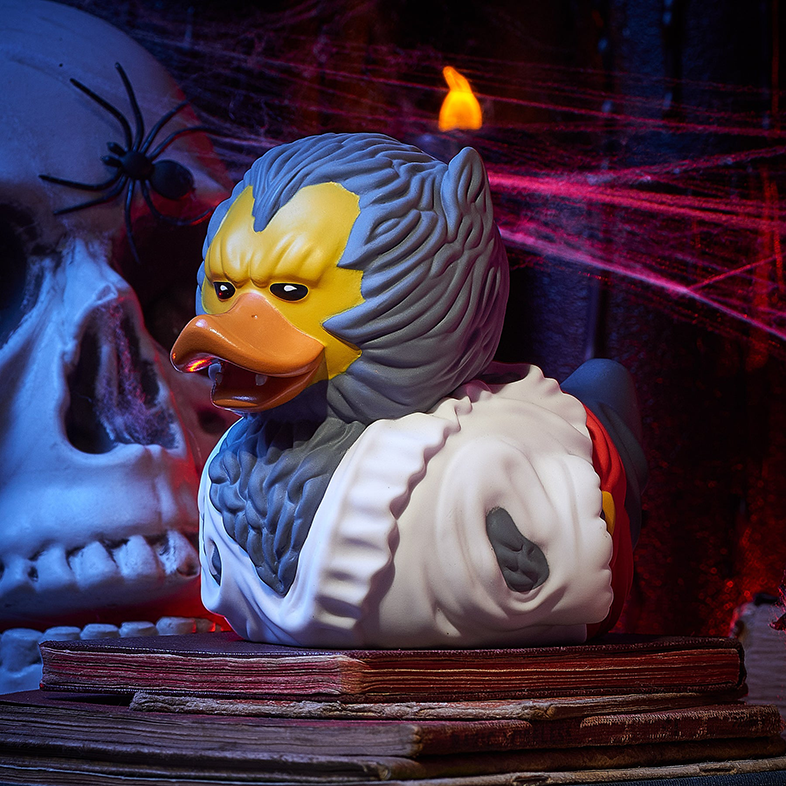 Halloween témájú gumikacsa, koponyával, pókkal és könyvvel a háttérben, narancssárga fénnyel megvilágítva.